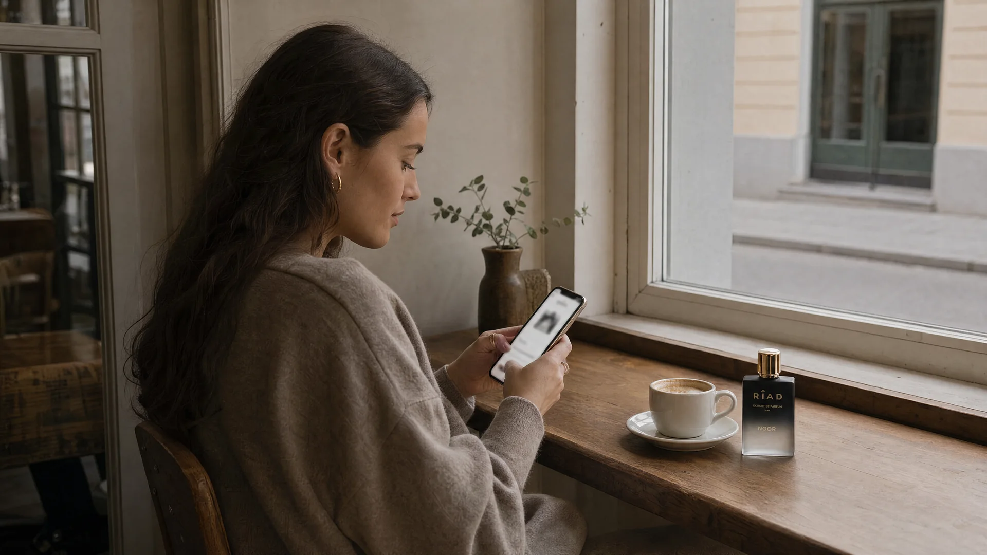 Ung kvinna vid ett fönsterbord på ett kafé scrollar på mobilen, Riads Noor står vid kaffekoppen
